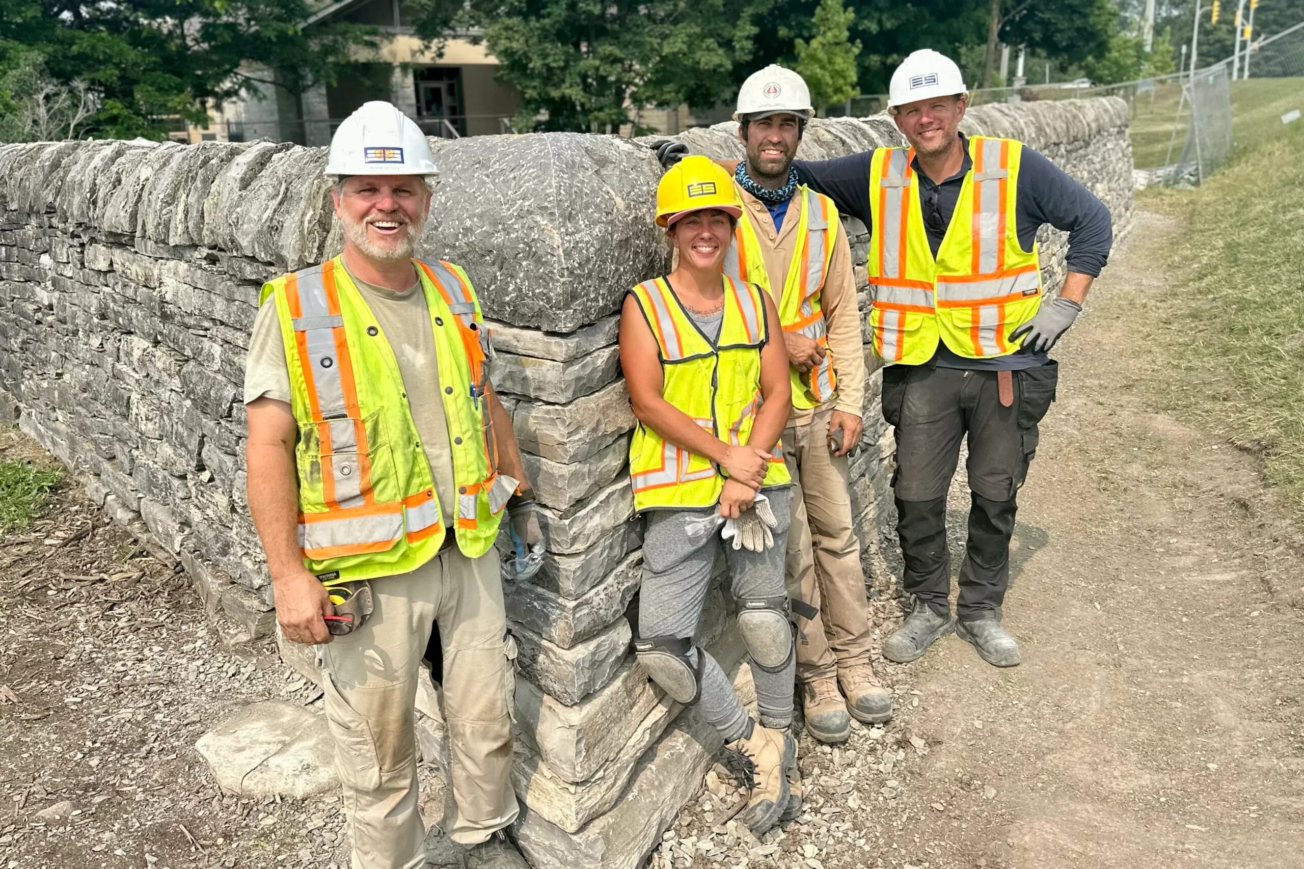 Four wallers in hard hats and hi-vis at the finished corner of the rebuilt Kingston wall, the large shaped corner stone and its cope beside them.