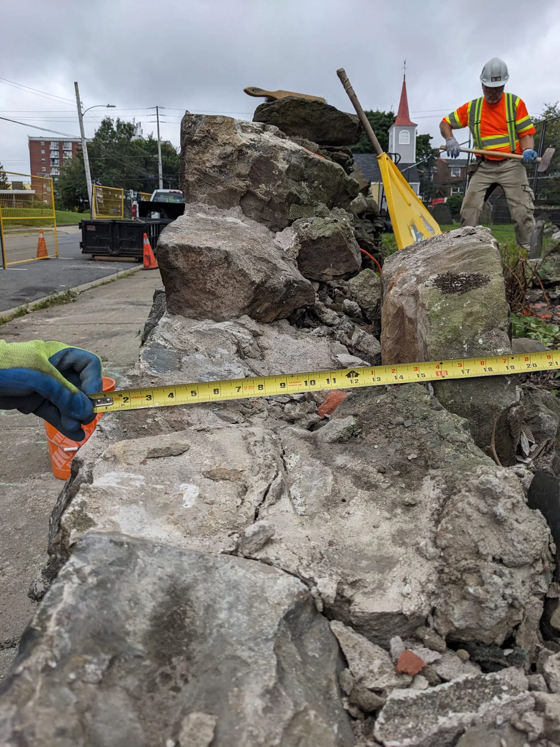 A waller measuring a dismantled section of heritage dry stone wall with a tape, a second waller working behind, and a church steeple in the distance.