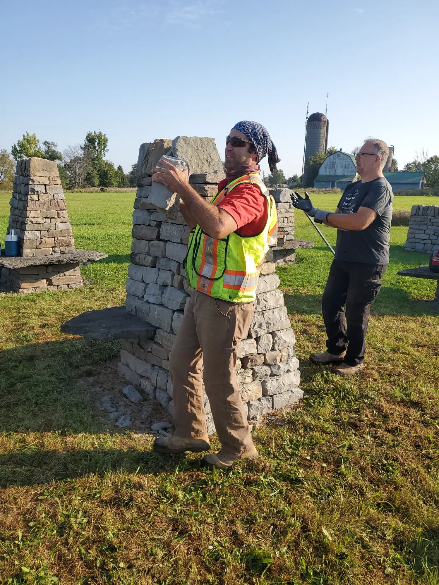 A waller in a hi-vis vest setting a stone into the rebuilt solar alignment pillar, a second waller behind him, and a barn and silo on the horizon.